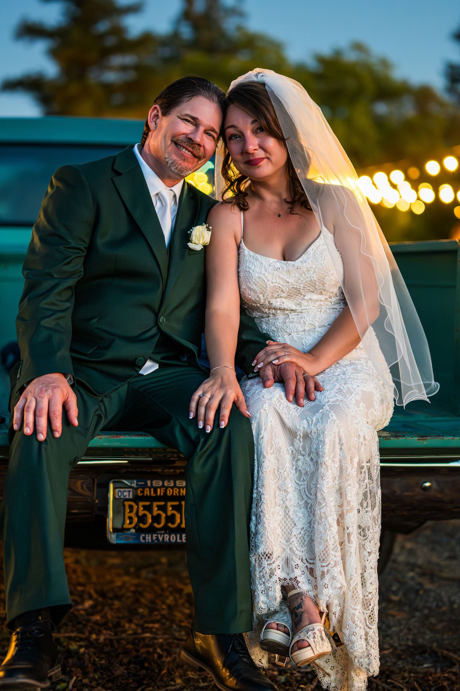 A romantic wedding couple sits together on a vintage car showing their Nevada license plate at sunset.