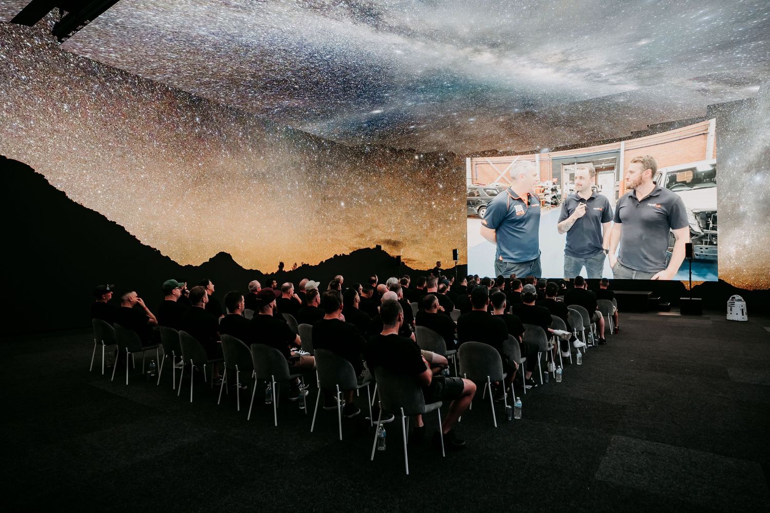 Silhouettes of people seated in rows watching an outdoor movie screening at dusk with a large projection screen visible.