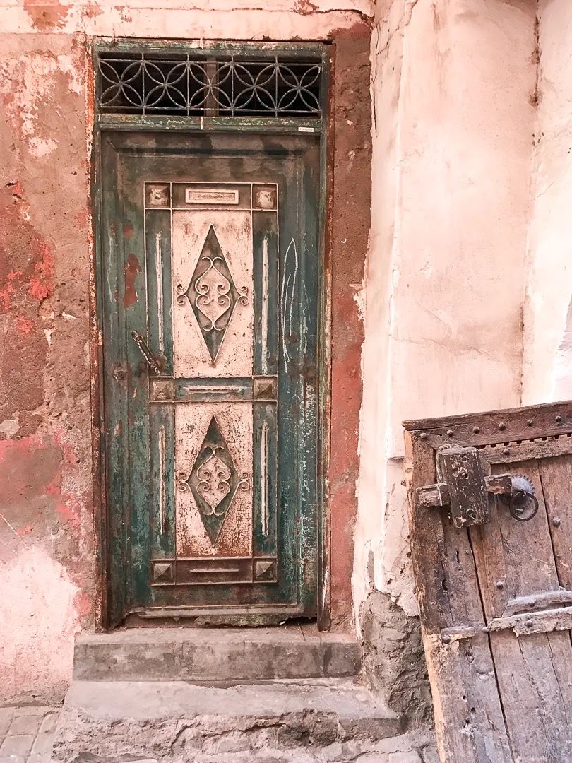 A weathered green Moroccan door with intricate carvings set in a soft pink plaster wall.