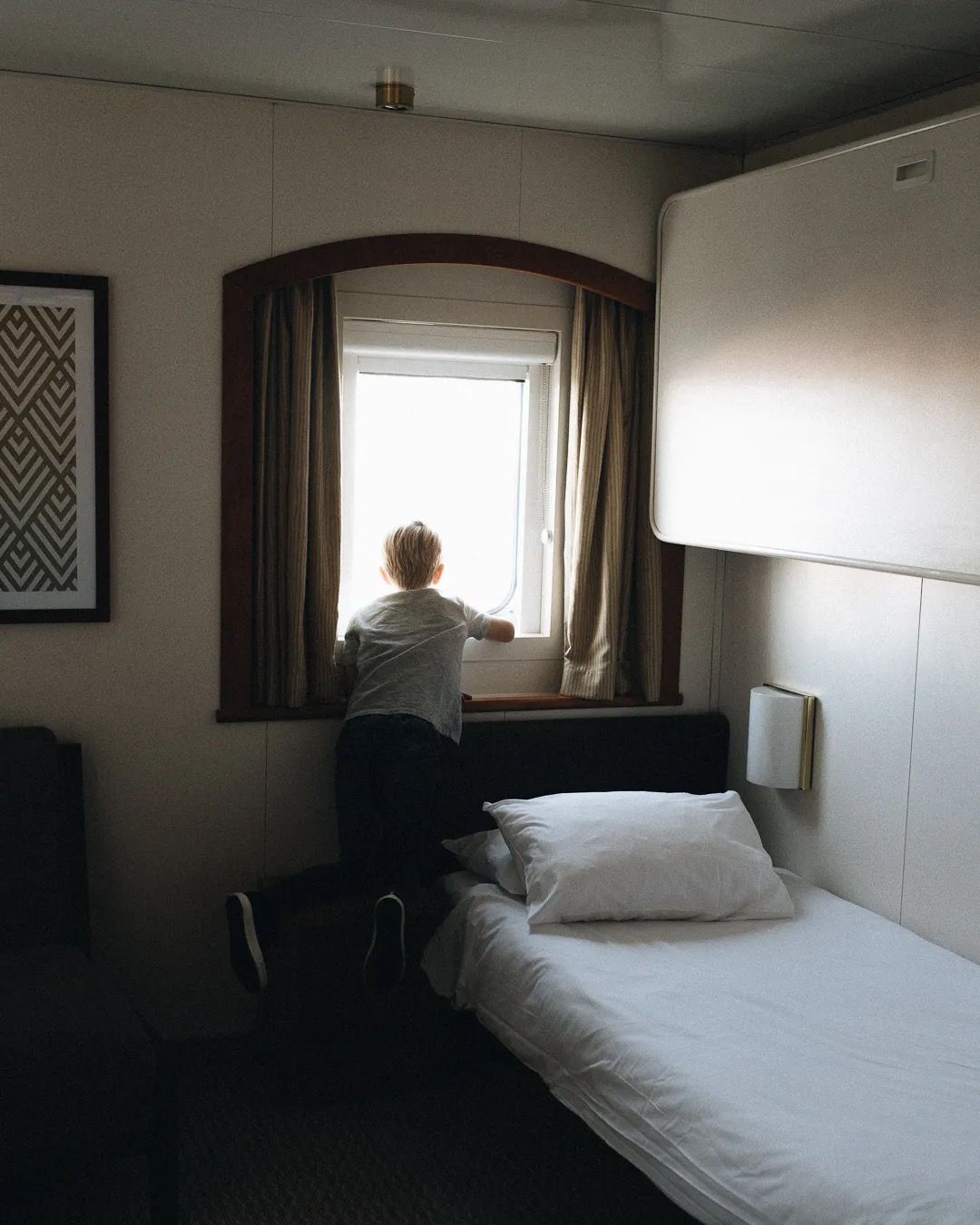 Child sitting on a bed near a large window in a quiet hotel room.