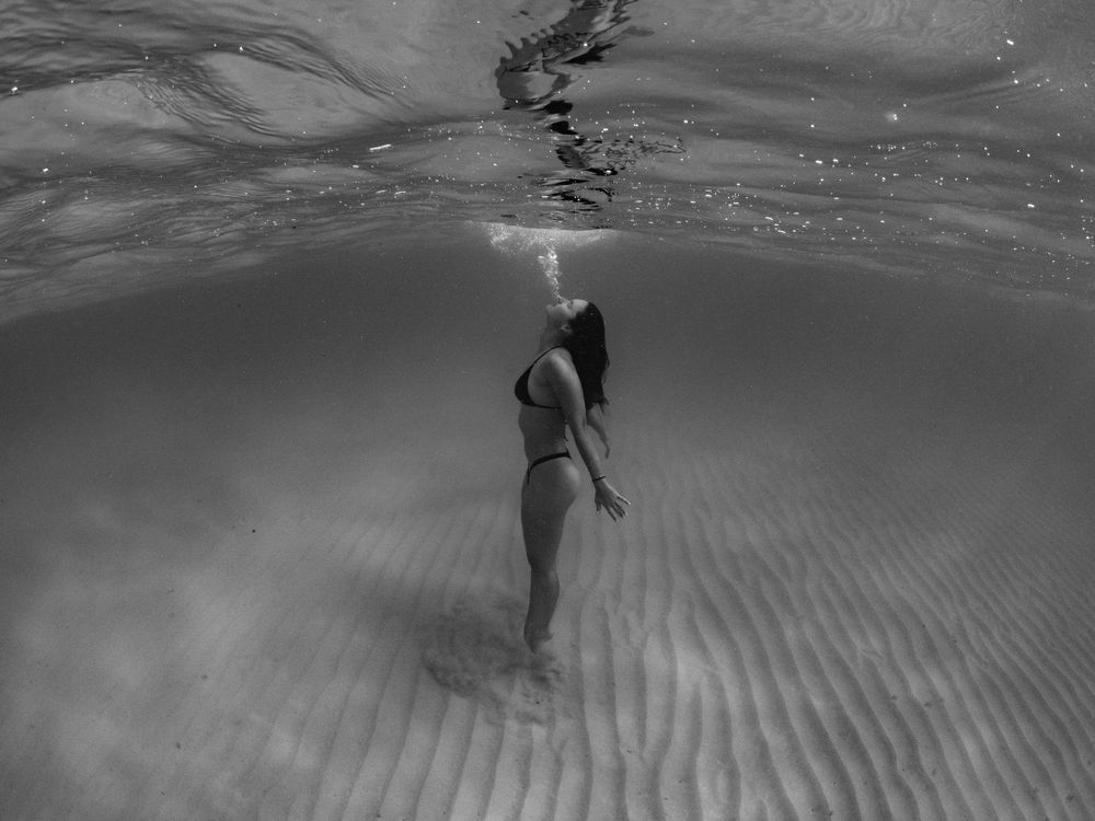 Monochrome underwater image showing a swimmer suspended vertically beneath the water's surface.