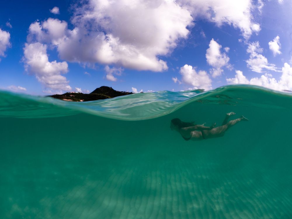 Split-view underwater photo showing a swimmer beneath turquoise waves with puffy white clouds visible above the surface.