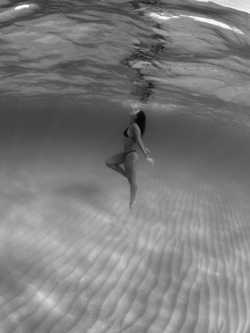 Black and white underwater photo of a swimmer floating gracefully above rippled sandy seafloor.