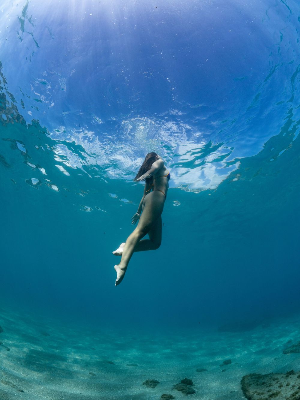 Underwater photograph of a swimmer descending through clear blue waters above a sandy ocean floor.