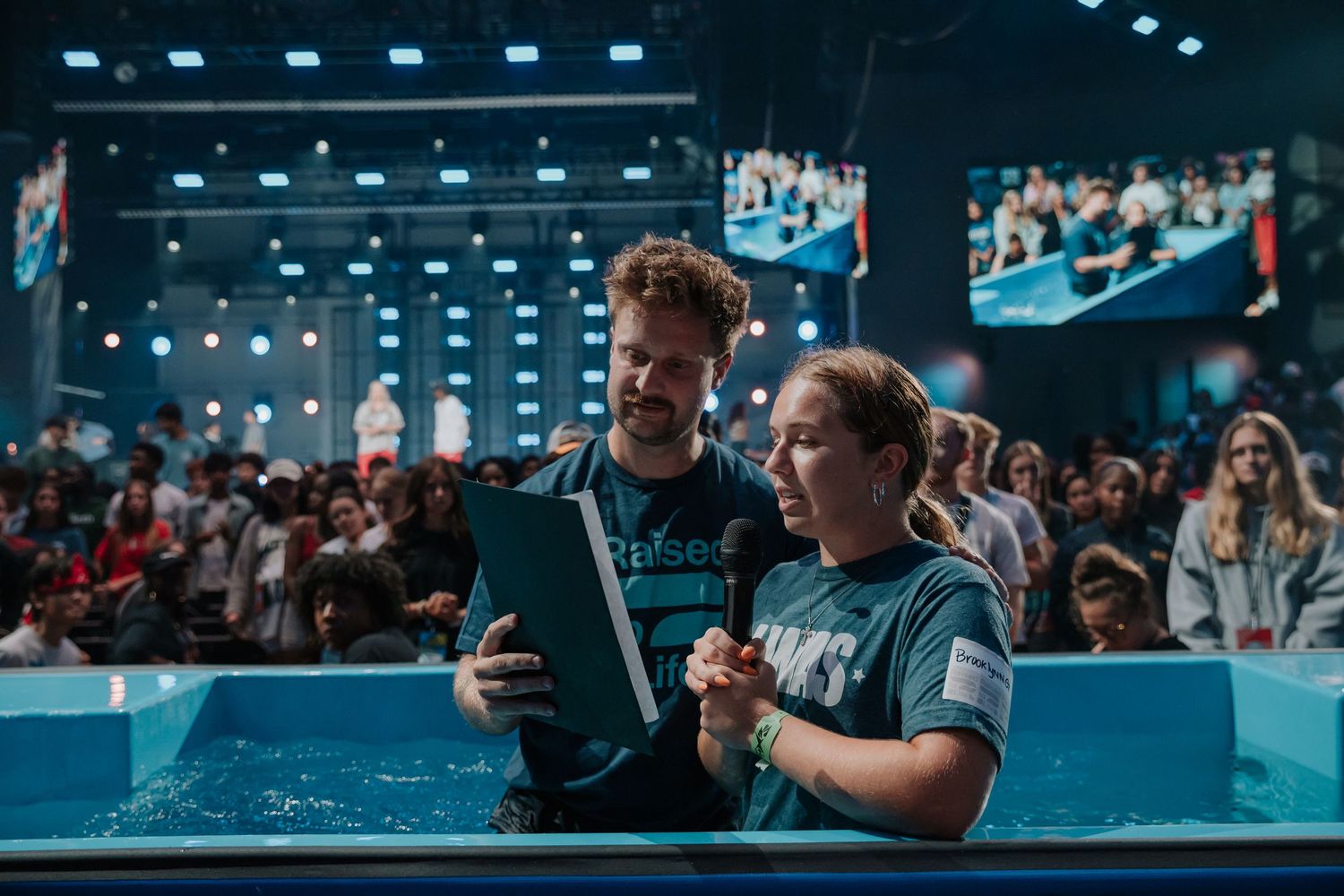 Pastor reads from a tablet with a young woman before her baptism in a large church baptismal pool.