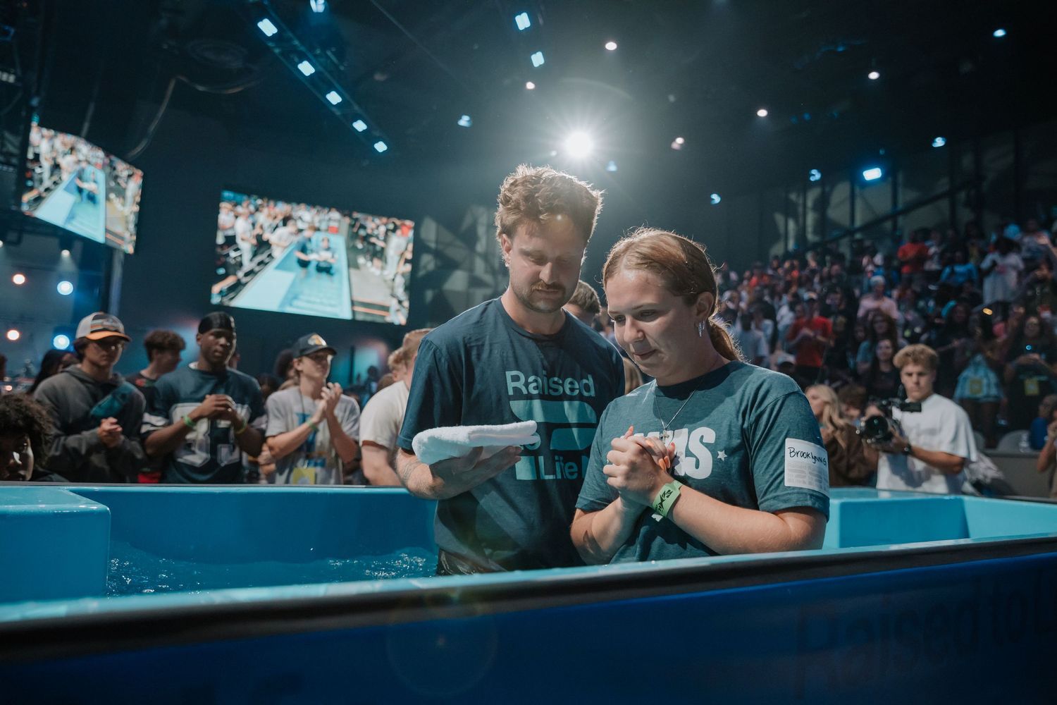 Pastor and young woman review baptism scripture together standing in a church baptismal pool before a crowd.