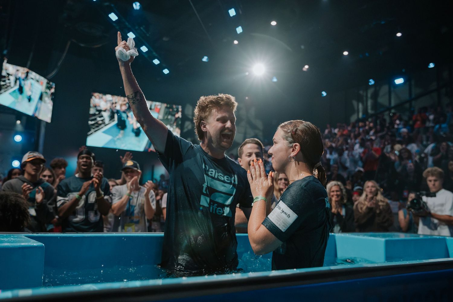 Joyful pastor raises finger to sky celebrating with a newly baptized young woman in a church baptismal pool.