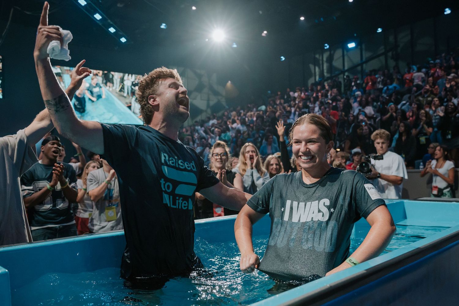 Two people celebrate joyfully in a baptismal pool wearing I Was shirts before a cheering congregation.