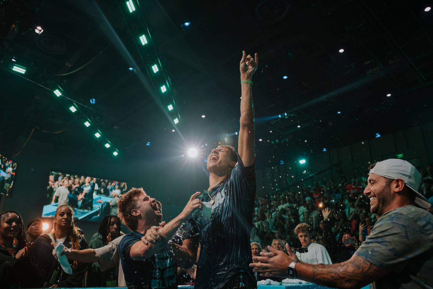 Newly baptized man raises arm in celebration surrounded by joyful friends in a large church auditorium.