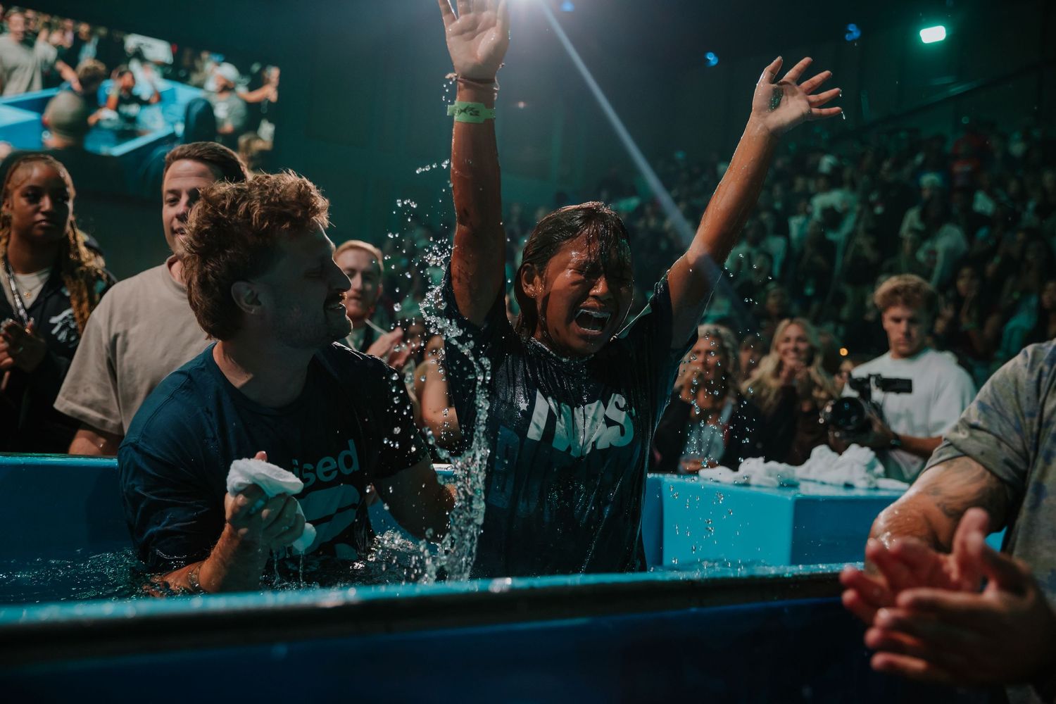 Teen raises arms in celebration after being baptized in water tank during large church service.