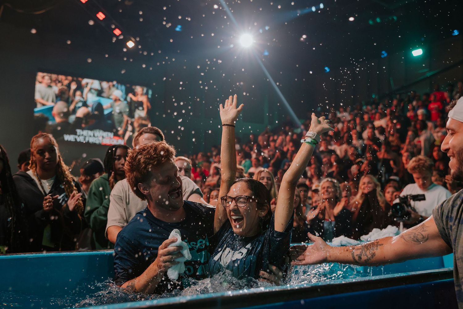Young woman joyfully raises hands during baptism ceremony in a crowded church auditorium with confetti.