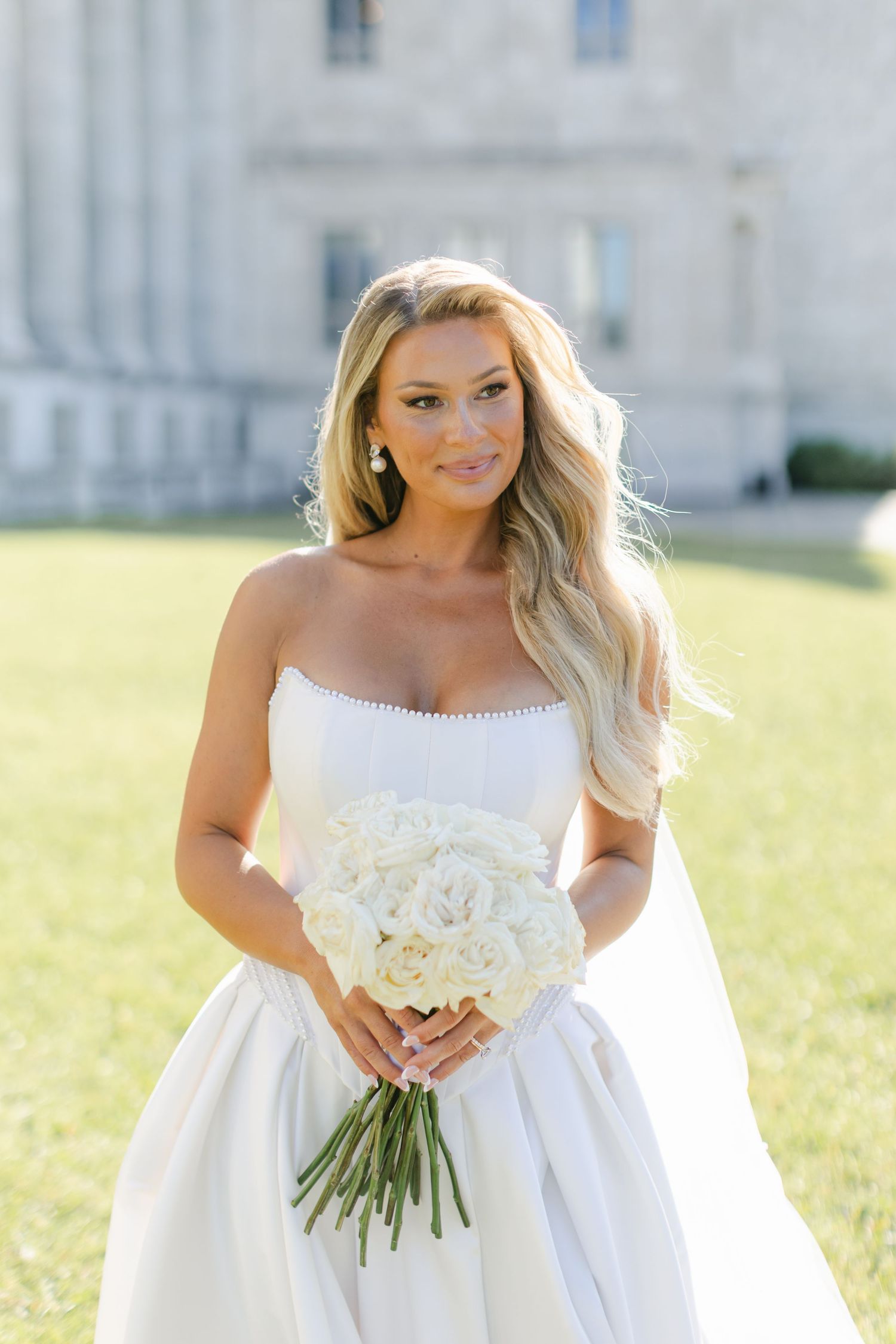 A bride in a white wedding dress holds a bouquet while standing in a sunny field near a grand building.