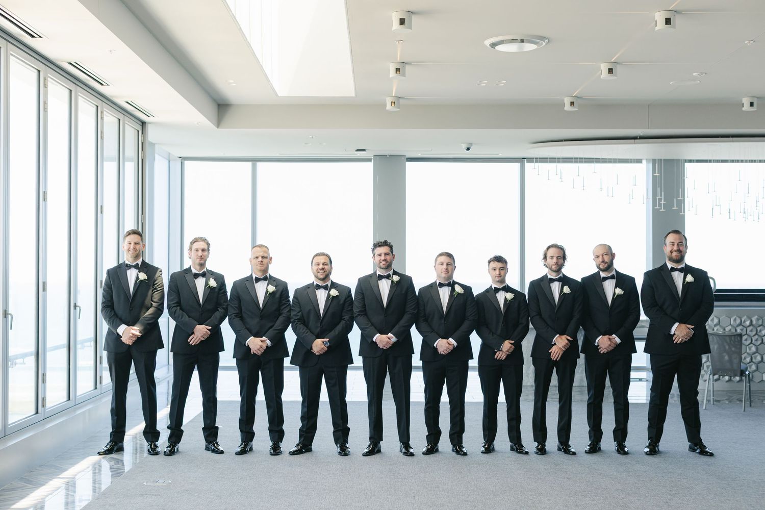 A row of groomsmen in black tuxedos and bow ties stand together against a white wall at a formal event.