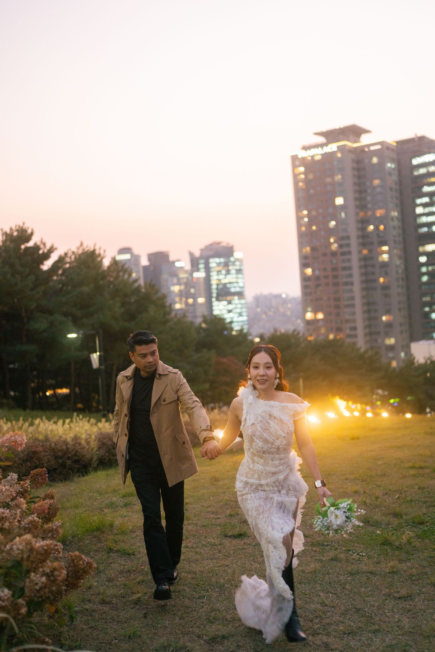 A couple runs through a park at sunset with city buildings and glowing lights in the background.
