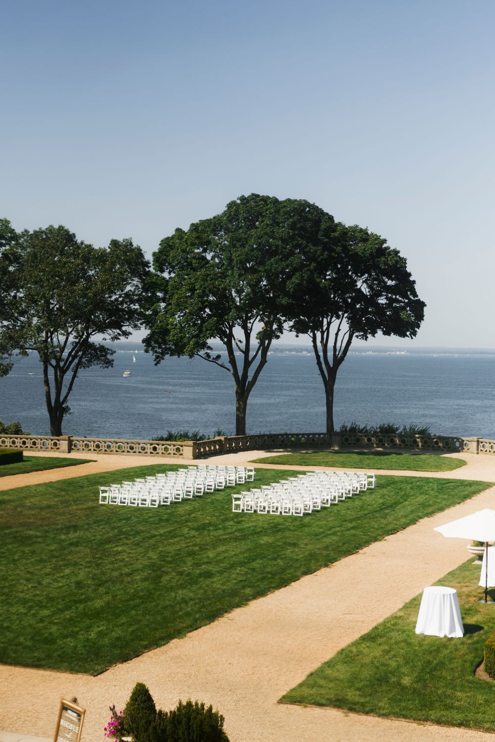 Outdoor wedding ceremony setup with white chairs on manicured lawn overlooking the ocean and trees.