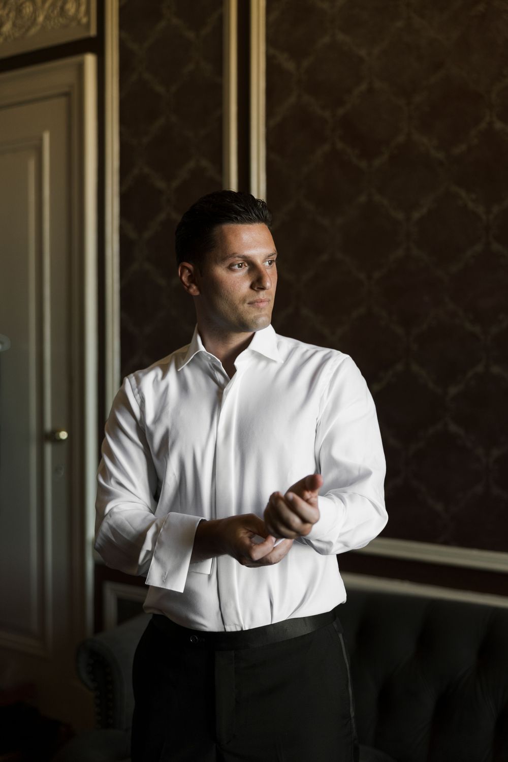 Groom in white dress shirt adjusting cufflinks in a dark elegant room with patterned wallpaper.