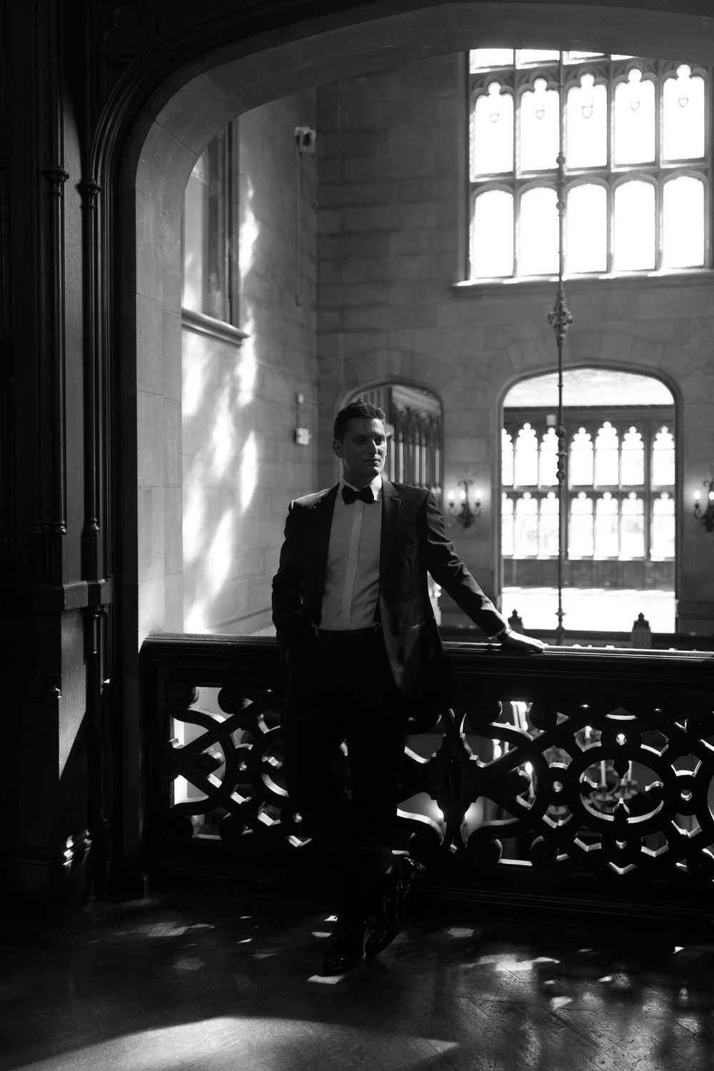 Groom in tuxedo leaning on ornate balcony railing in dramatic black and white gothic interior.