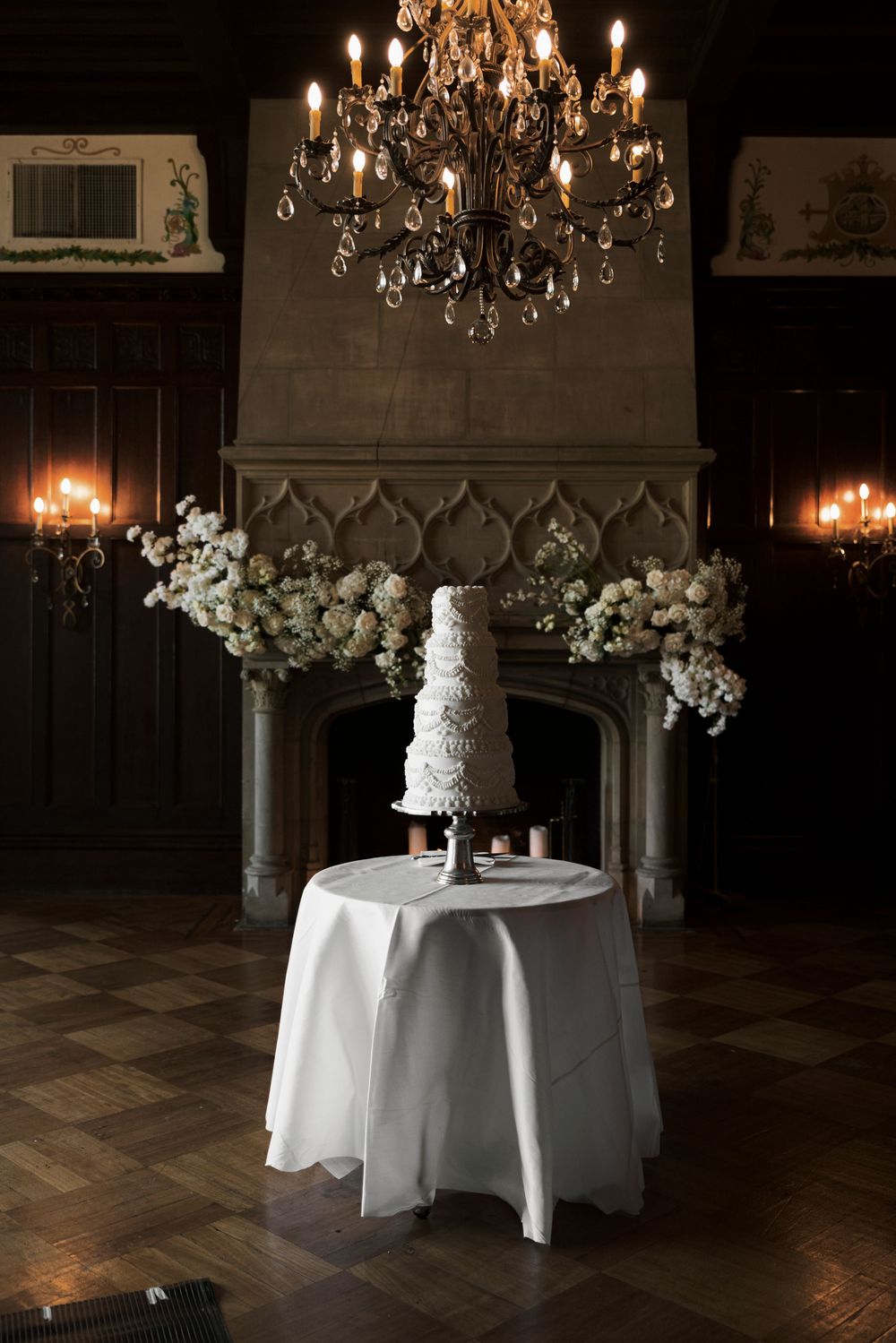 White tiered wedding cake on draped table before a gothic stone fireplace with floral arrangements and chandelier.