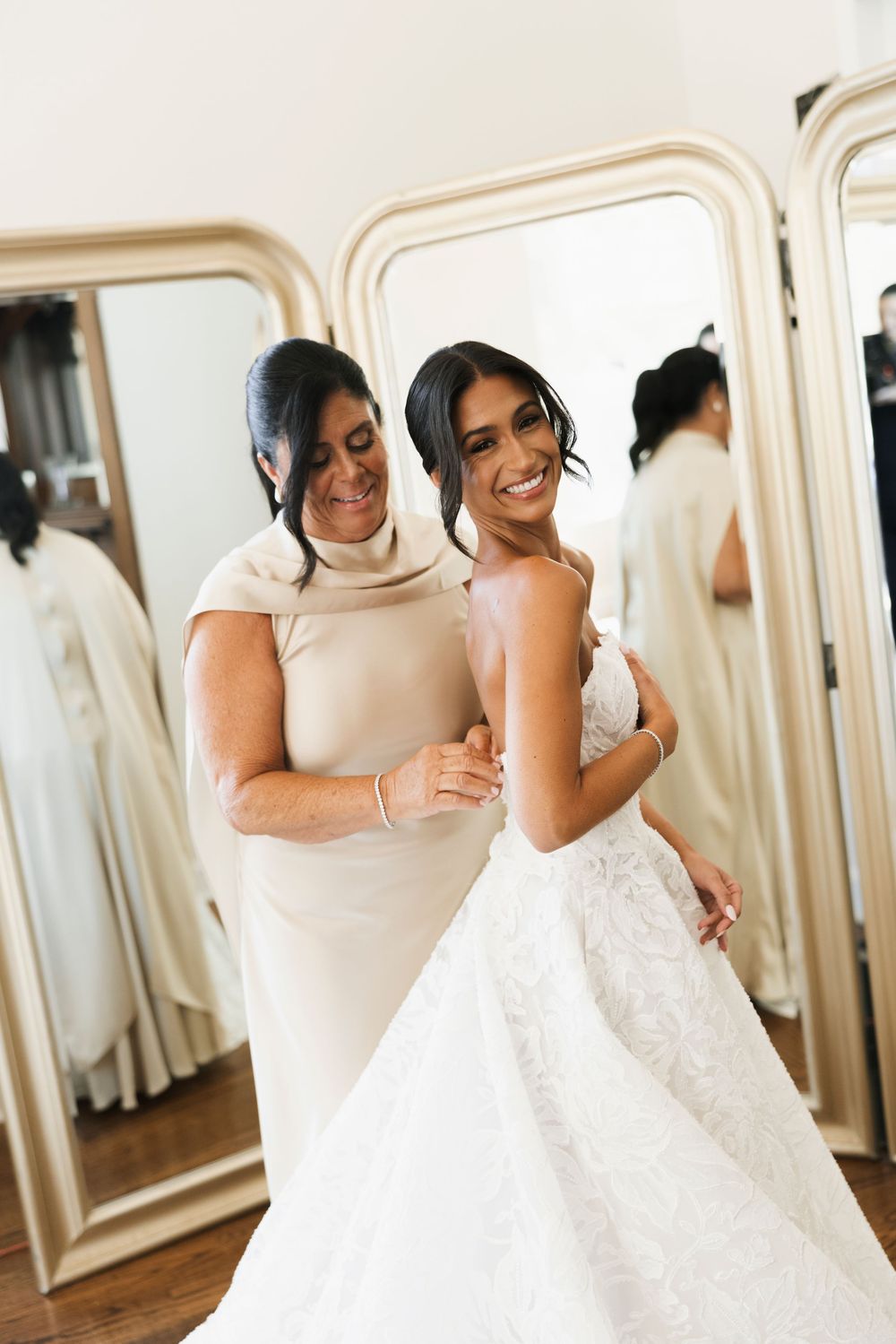 Smiling bride in strapless lace wedding gown being helped by her mother in front of gold-framed mirrors.