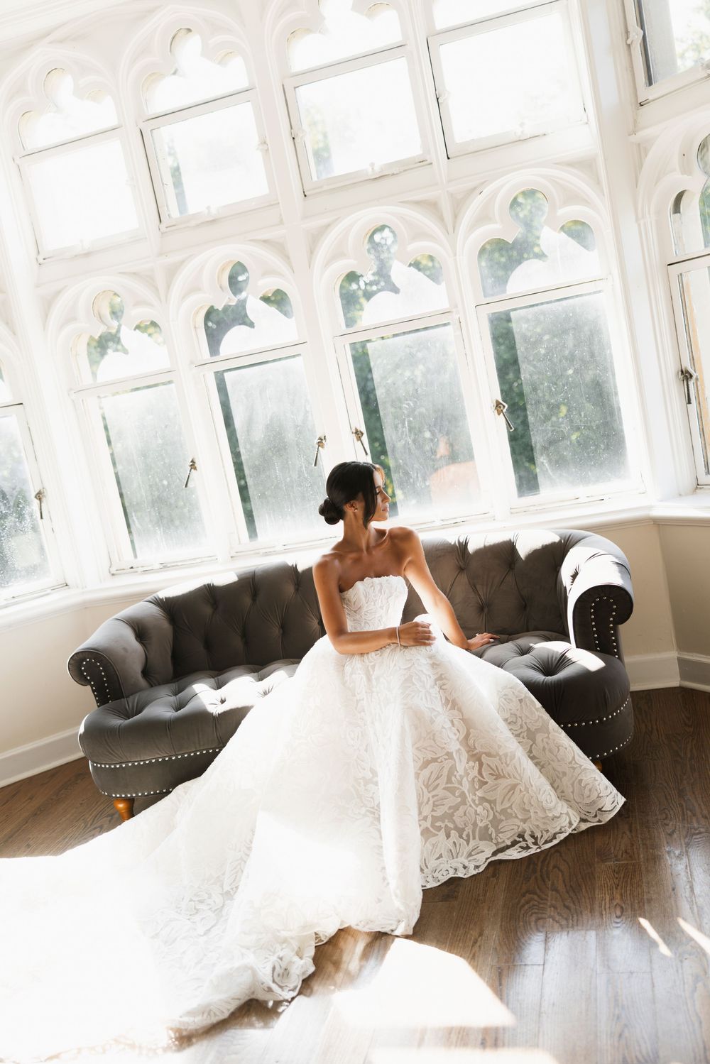 Bride in strapless lace ball gown with long train sitting on a tufted sofa before large gothic windows.