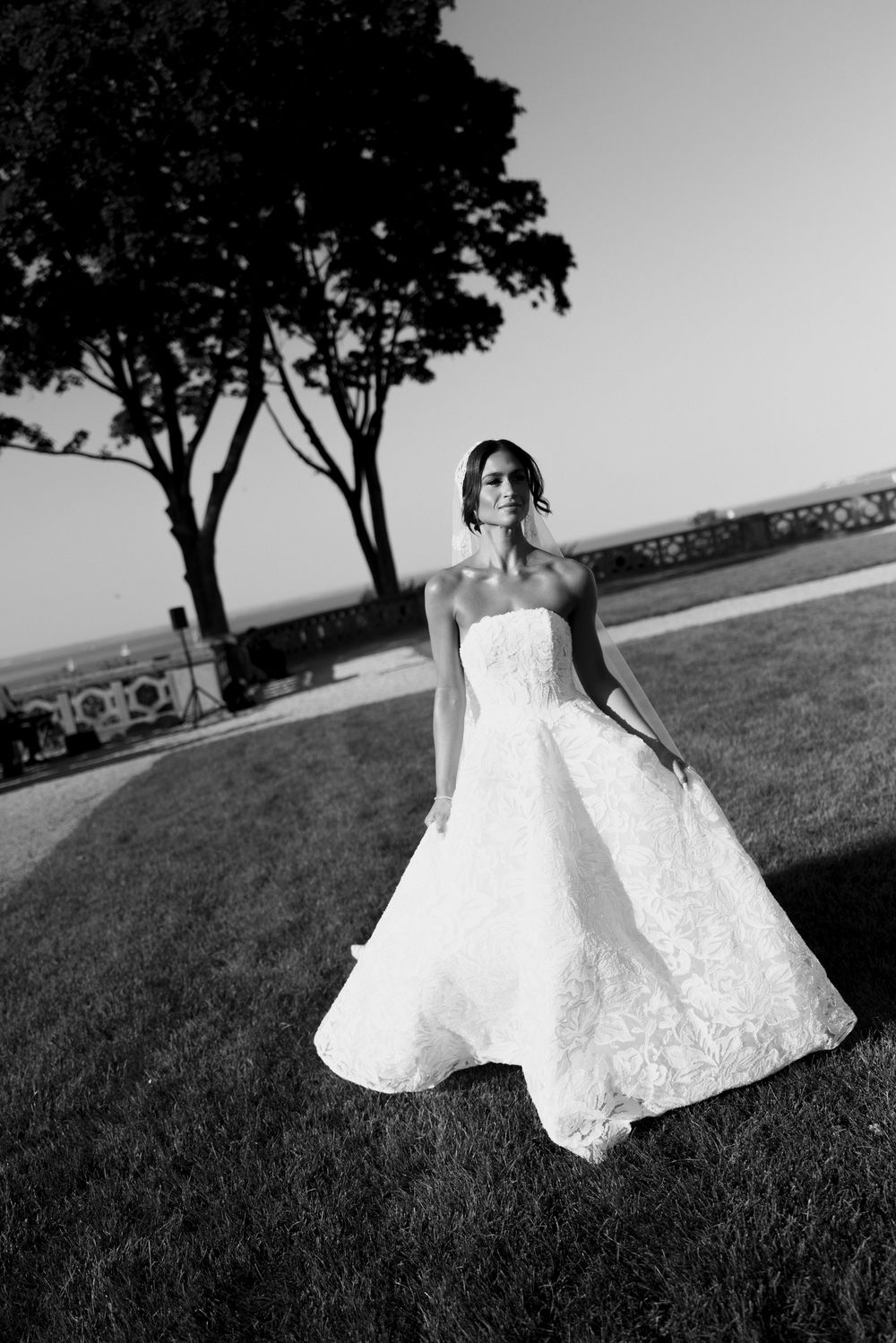 Bride in strapless lace ball gown wedding dress poses on green lawn with trees in black and white photo.