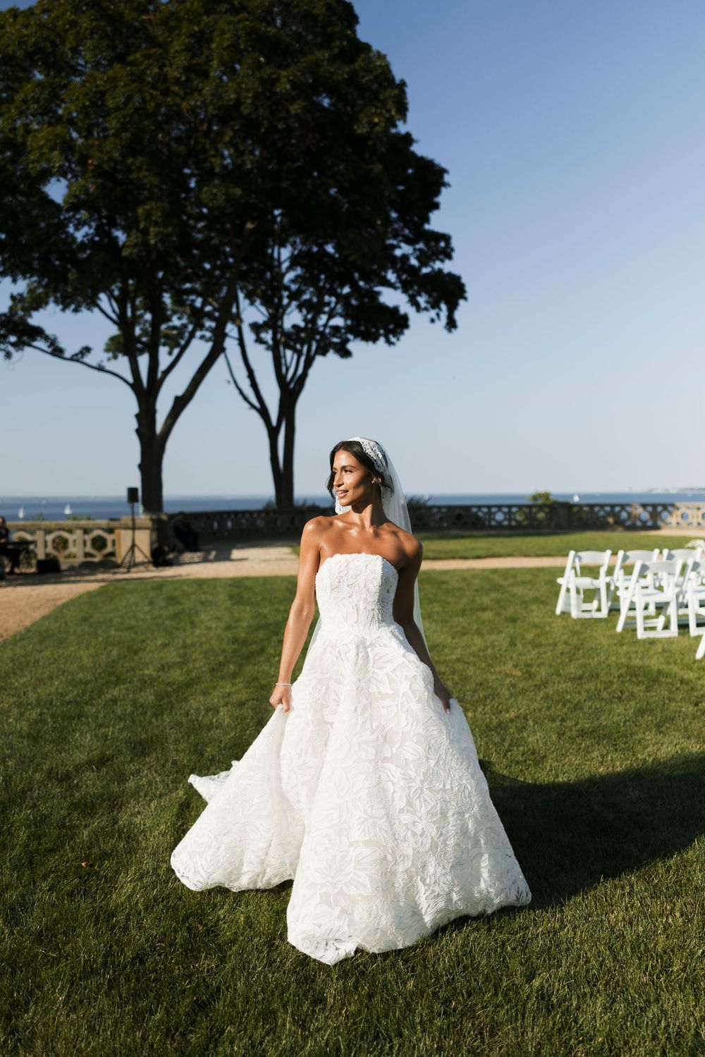 Bride in strapless lace ball gown and veil stands elegantly on lawn with white ceremony chairs behind her.