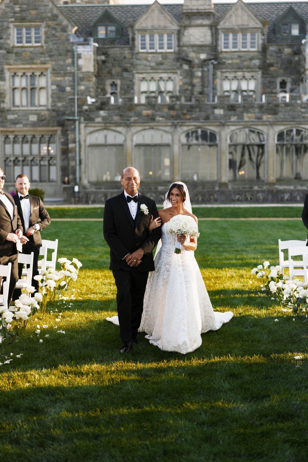 Father escorts radiant bride in lace gown down outdoor aisle at elegant castle estate wedding ceremony.