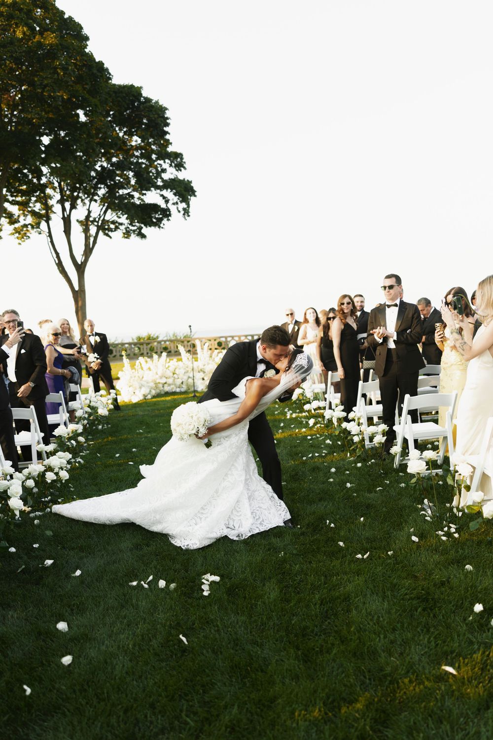 Groom dips bride in lace wedding gown for romantic kiss along rose petal-lined outdoor aisle.