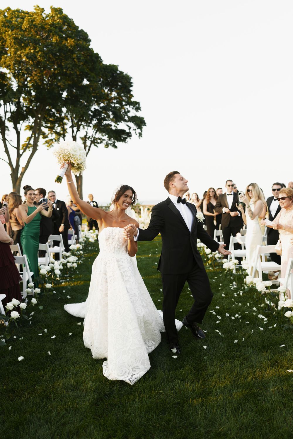Joyful bride raises bouquet as newlyweds celebrate walking back down outdoor ceremony aisle.