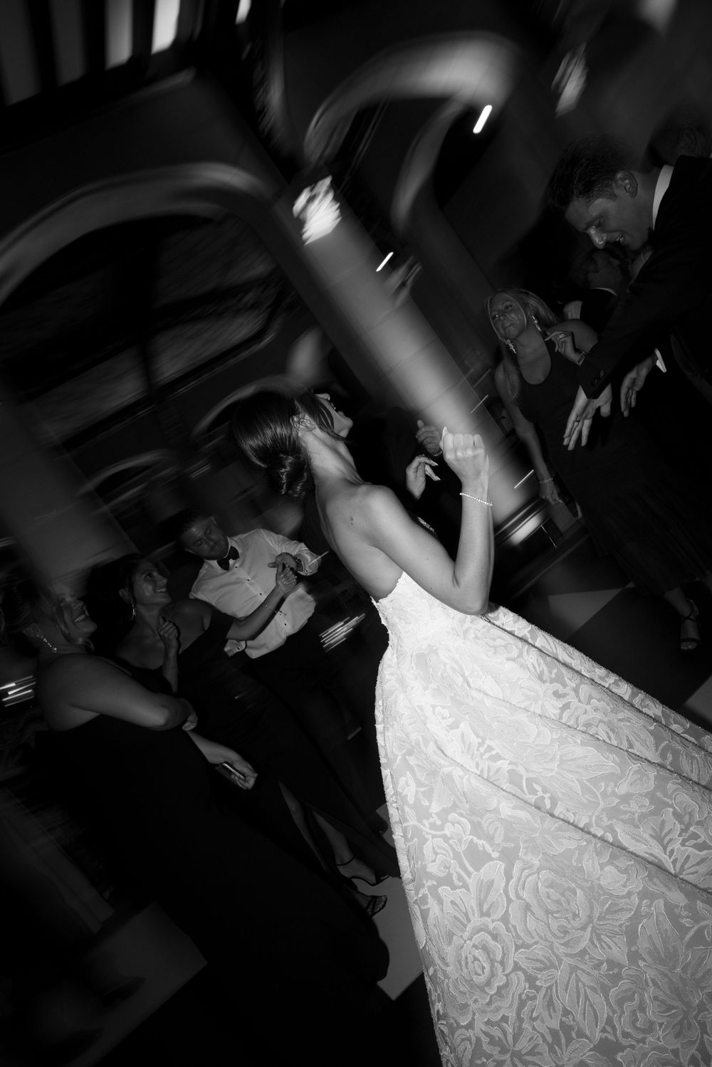 Black and white photo of a bride in lace gown dancing joyfully with guests at wedding reception.