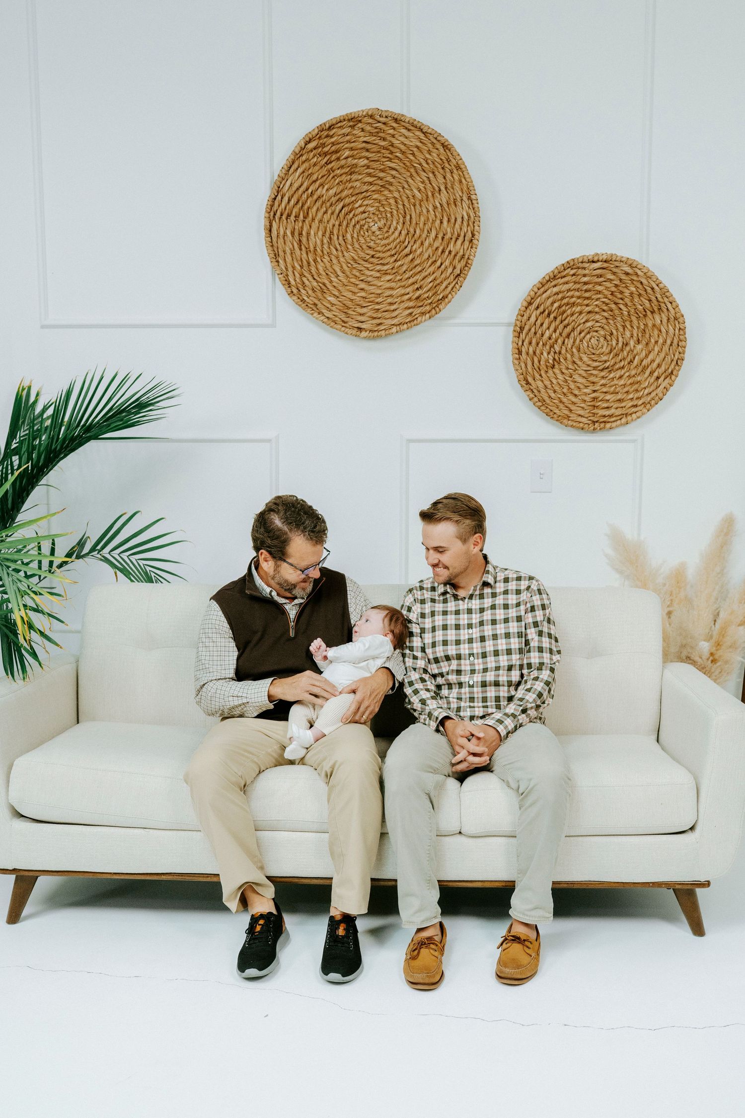 Two people sitting on a white couch with woven wall decor and palm plants in a minimalist setting.