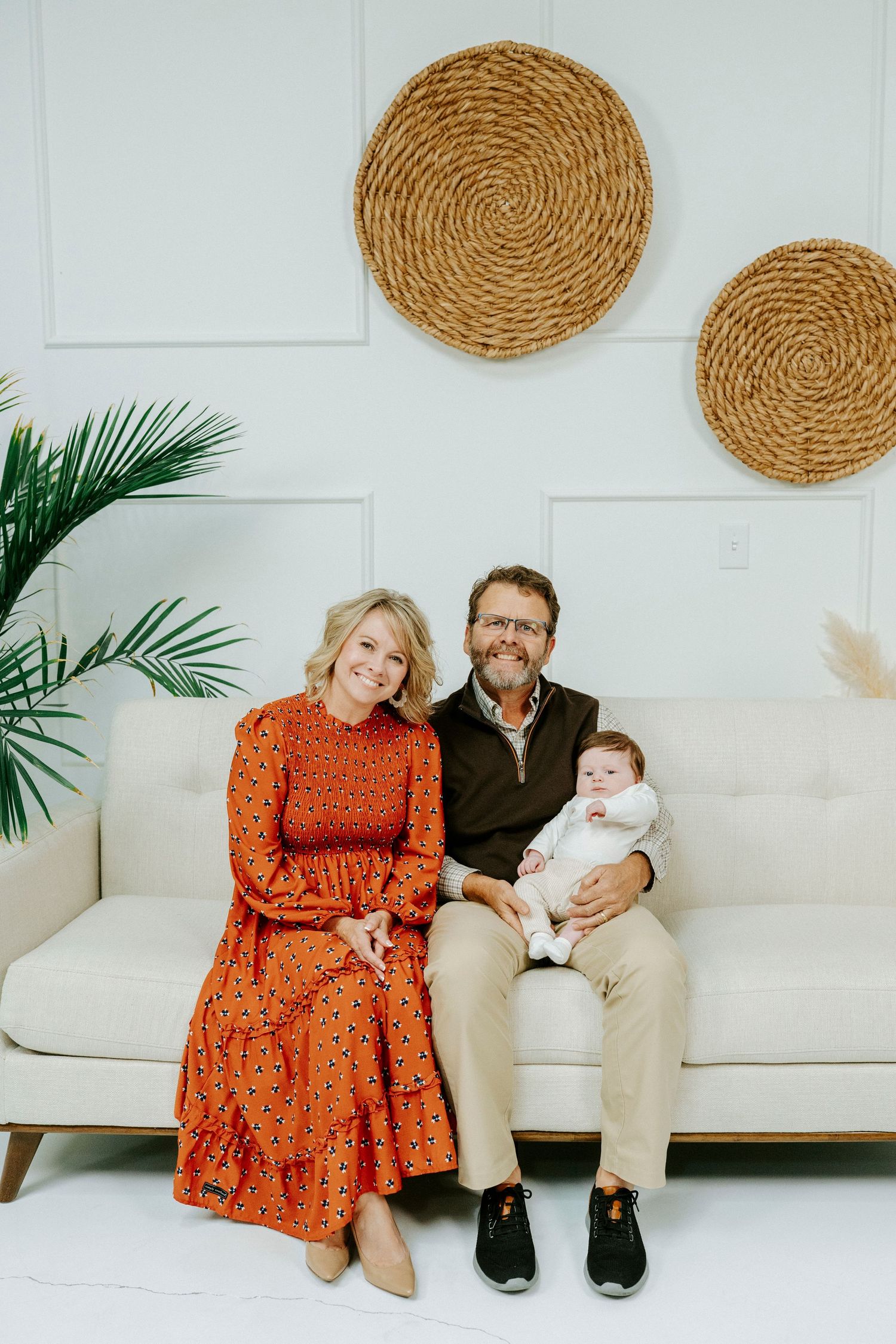 A family in coordinated orange and tan outfits sits together on a white couch with woven wall decor and palm leaves.