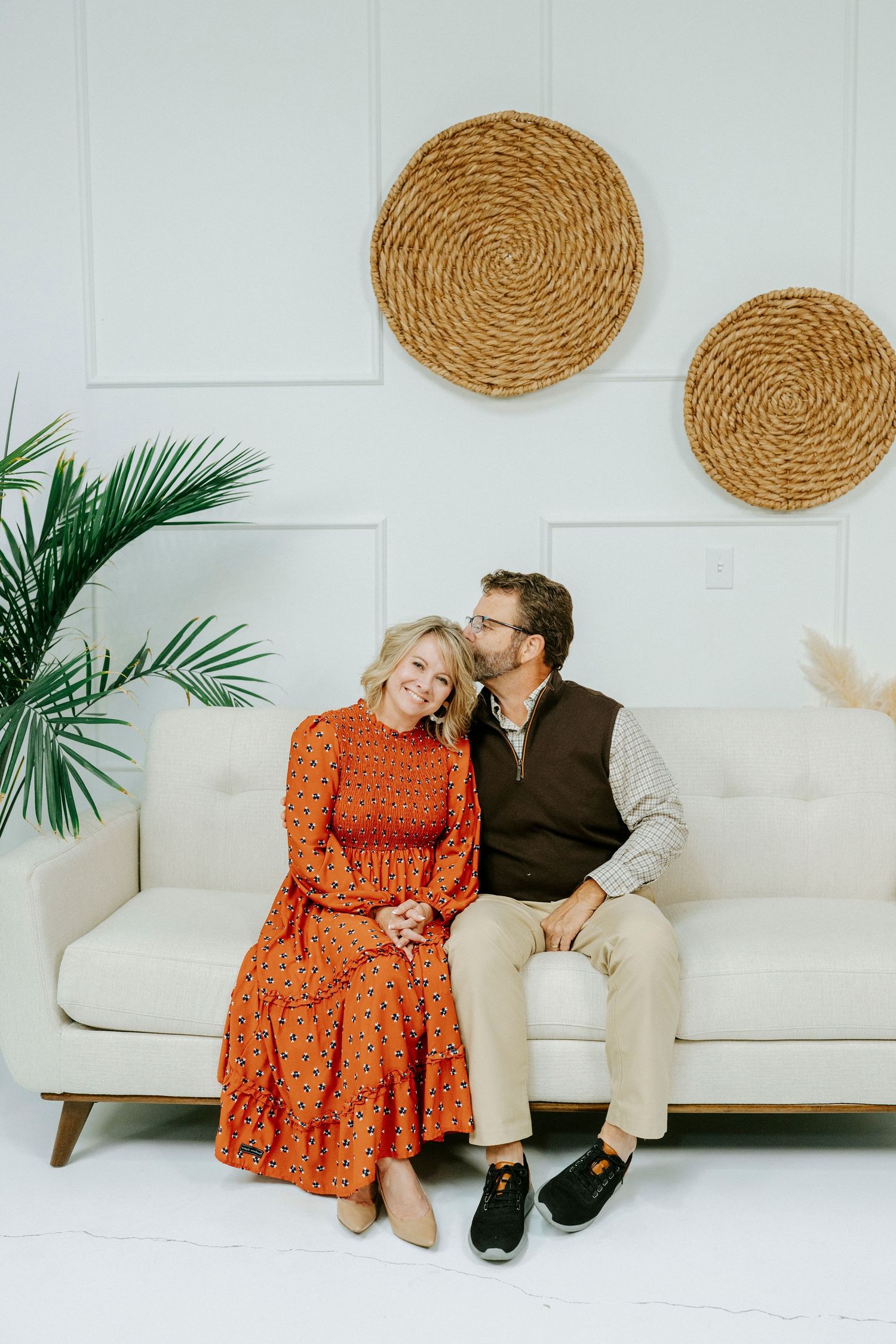 Couple sitting together on white couch with woven wall decor and palm plant in minimalist modern setting.
