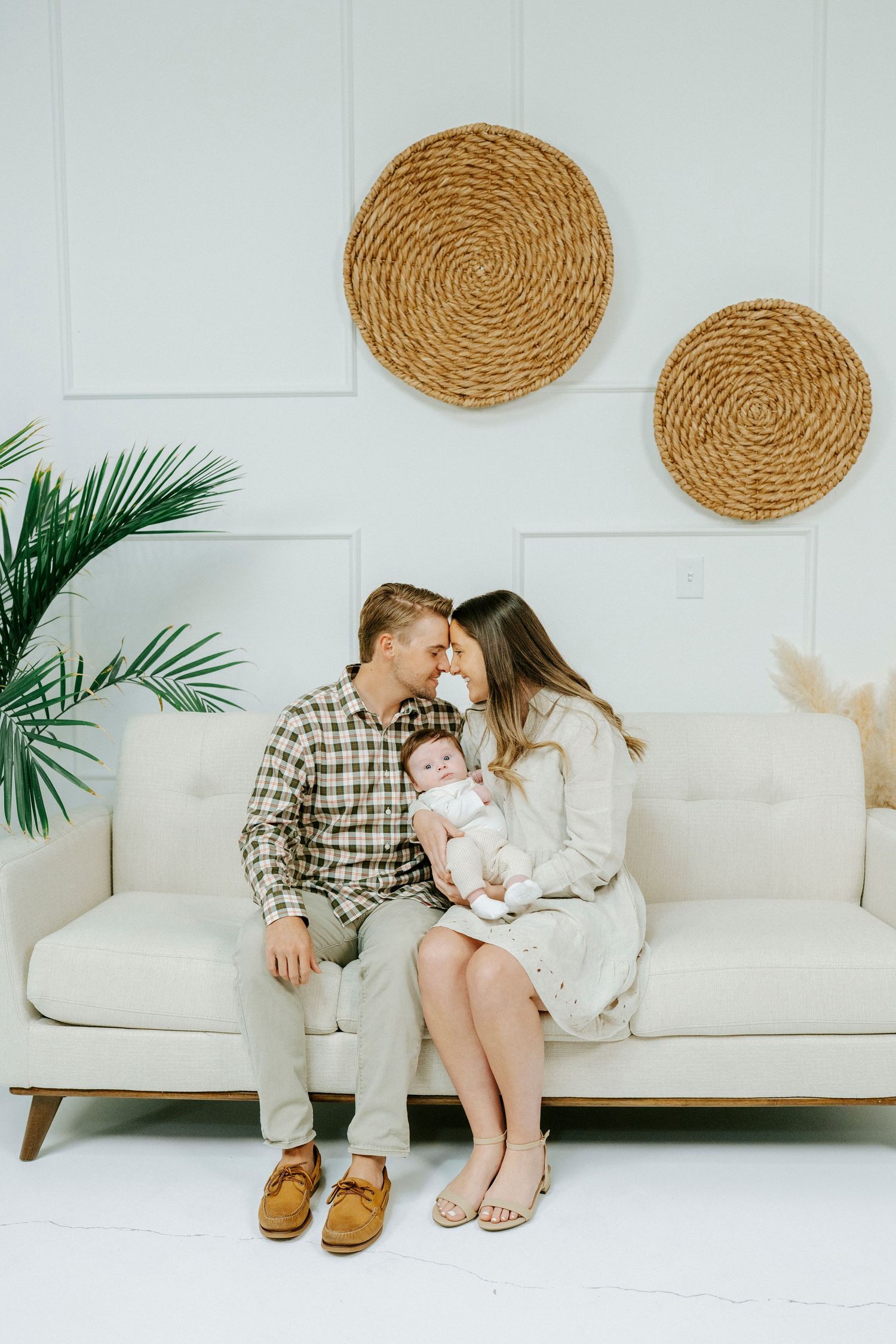 A family sits together on a white couch with circular woven wall decor and palm fronds in a bright modern setting.