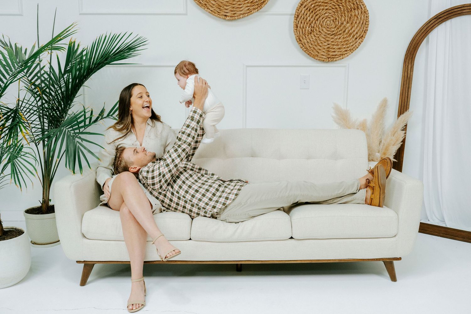 Cozy living room interior with woven wall baskets, white sofa, and tropical palm plants creating a modern boho atmosphere.