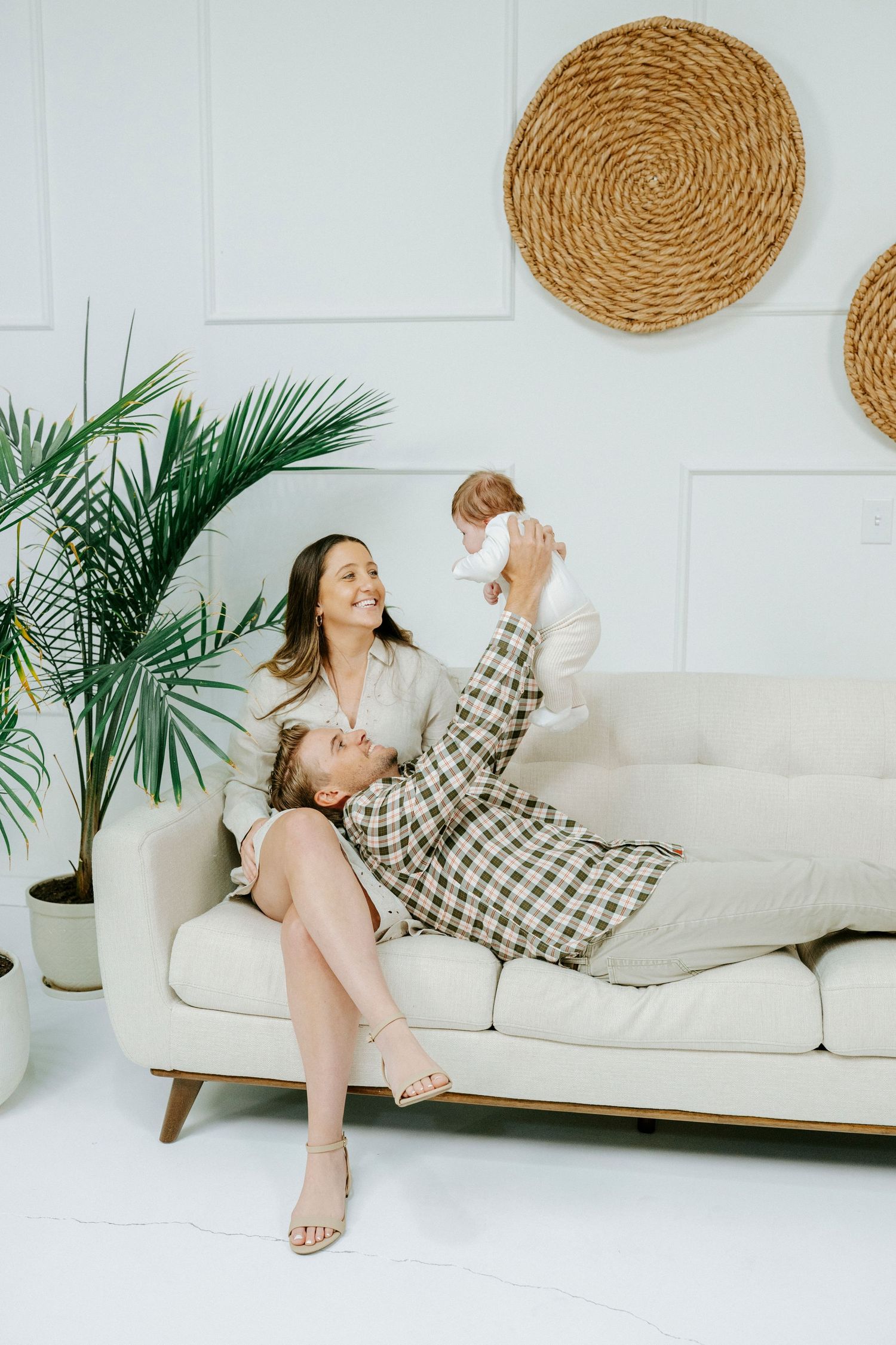 Two people sharing a playful moment on a white couch with decorative woven wall art and tropical plants in background.