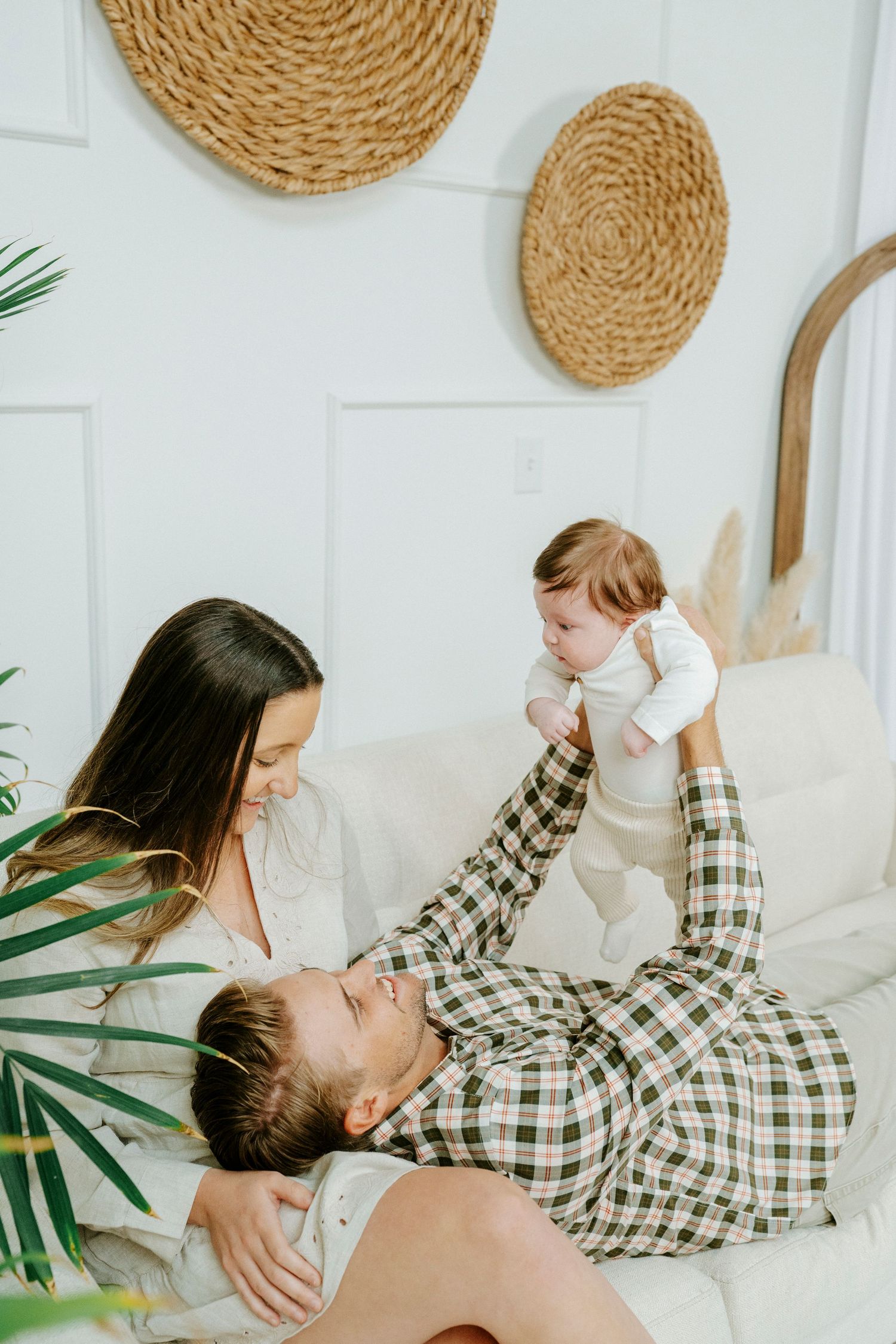 A couple lounges on a white couch with palm decor, sharing intimate moments in casual attire.