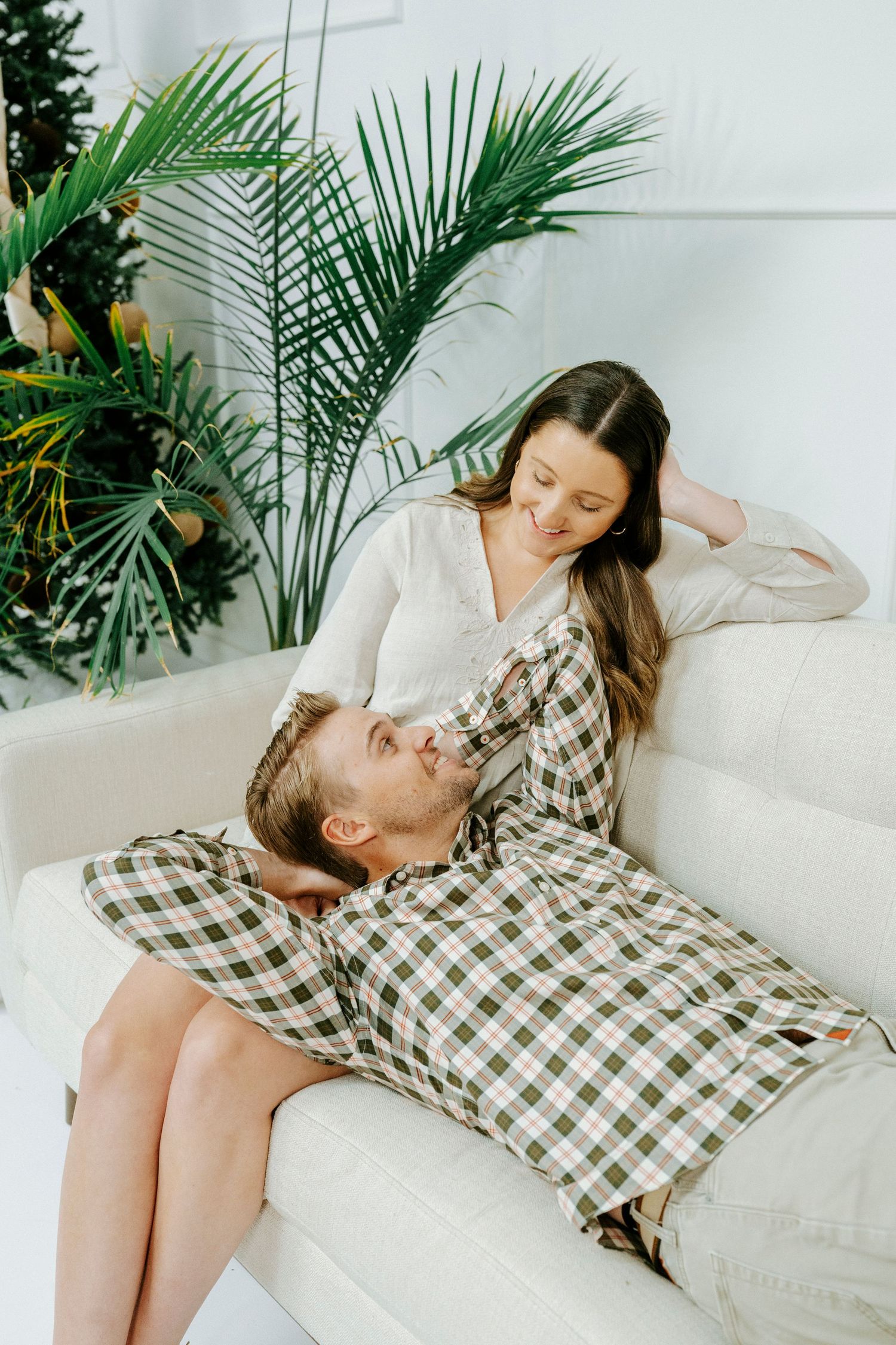A couple shares tender moments while relaxing on a white couch with tropical plants in the background.