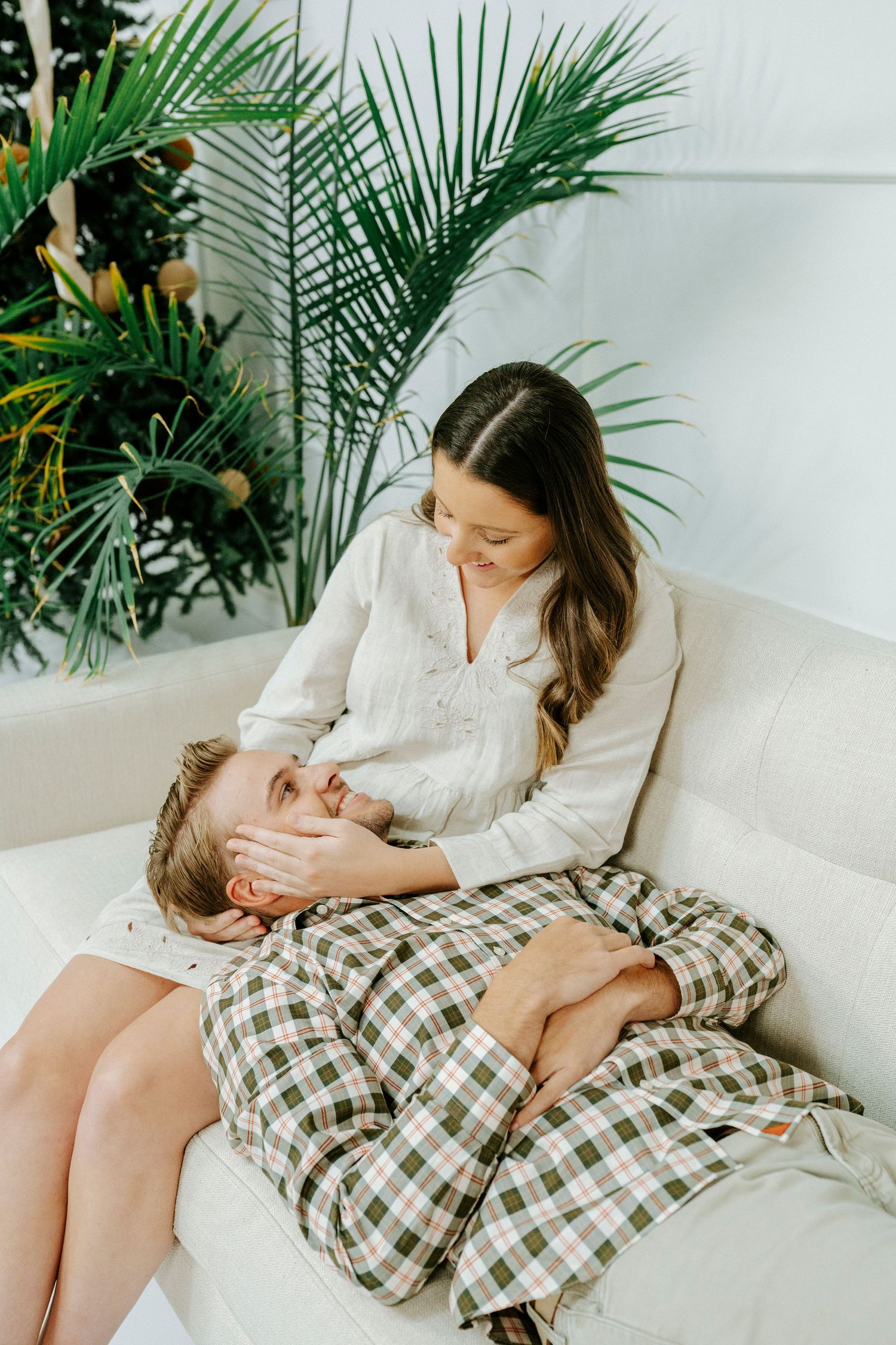 Couple relaxing together on white sofa with palm plant and natural decor elements in minimalist setting.
