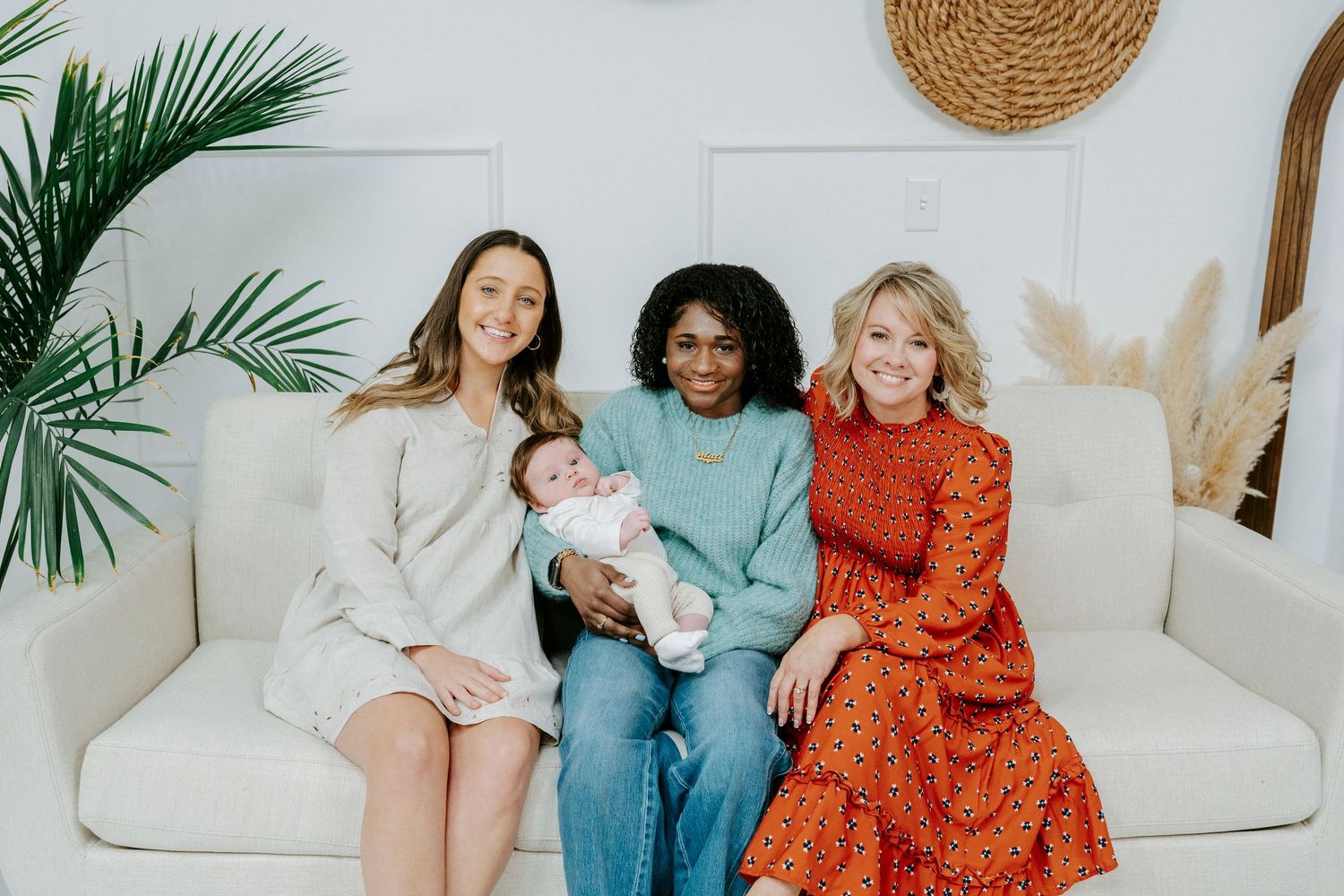 Three friends sit together on a white couch with a palm plant and boho decor in the background.