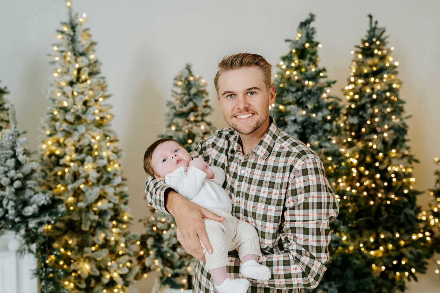 A festive holiday portrait in front of three illuminated Christmas trees with warm white lights.