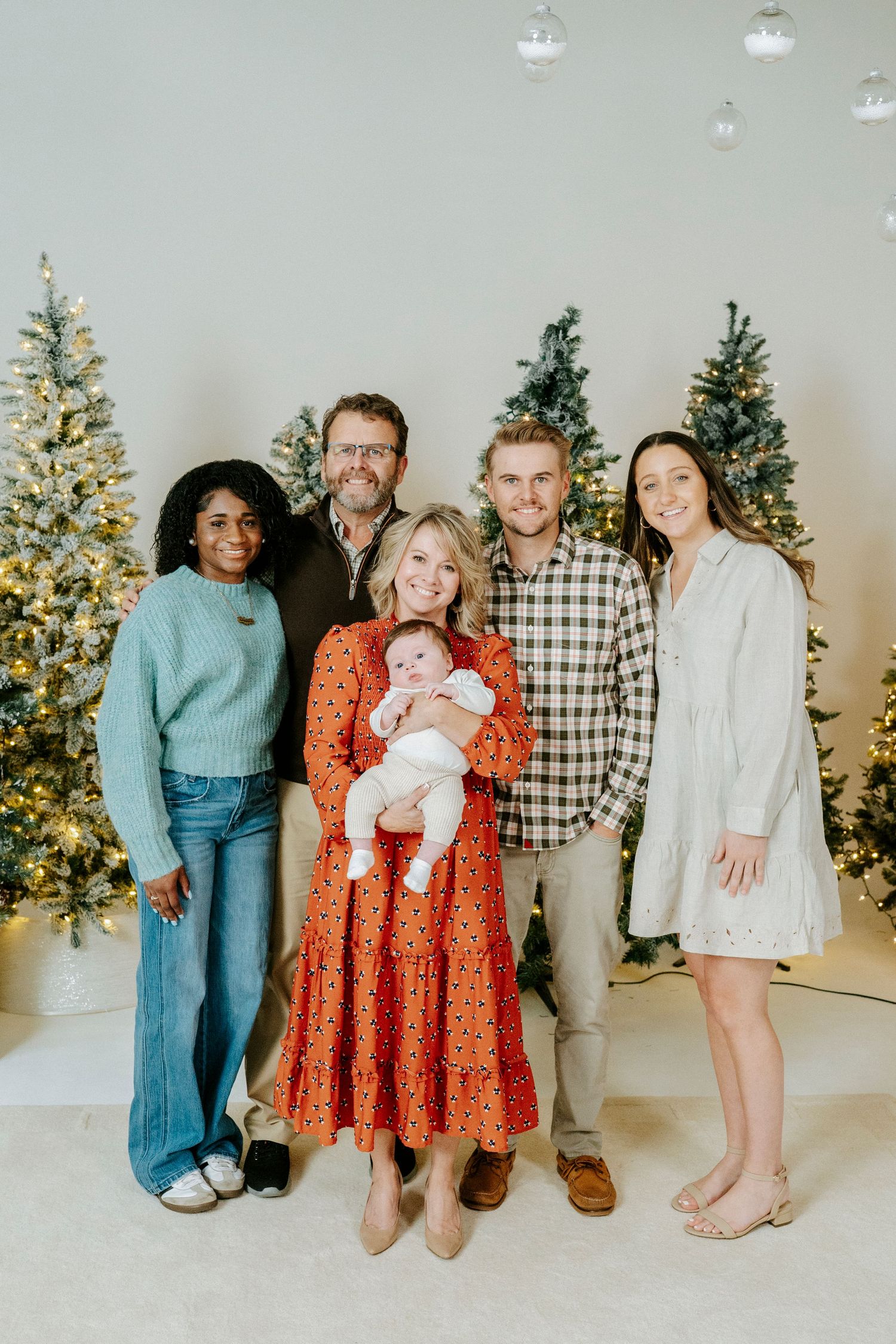A family poses together in front of decorated Christmas trees in a bright studio setting with white walls and floor.