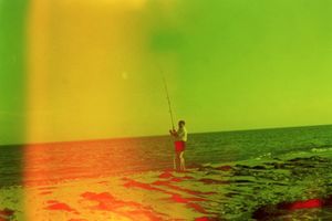 A silhouetted figure fishing on a beach at sunset with vibrant orange and green light effects.