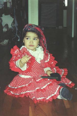 A small child wearing a red and white polka dot dress with ruffles sits on wooden flooring.