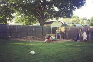 Large backyard with wooden fence and scattered play equipment under shady trees.