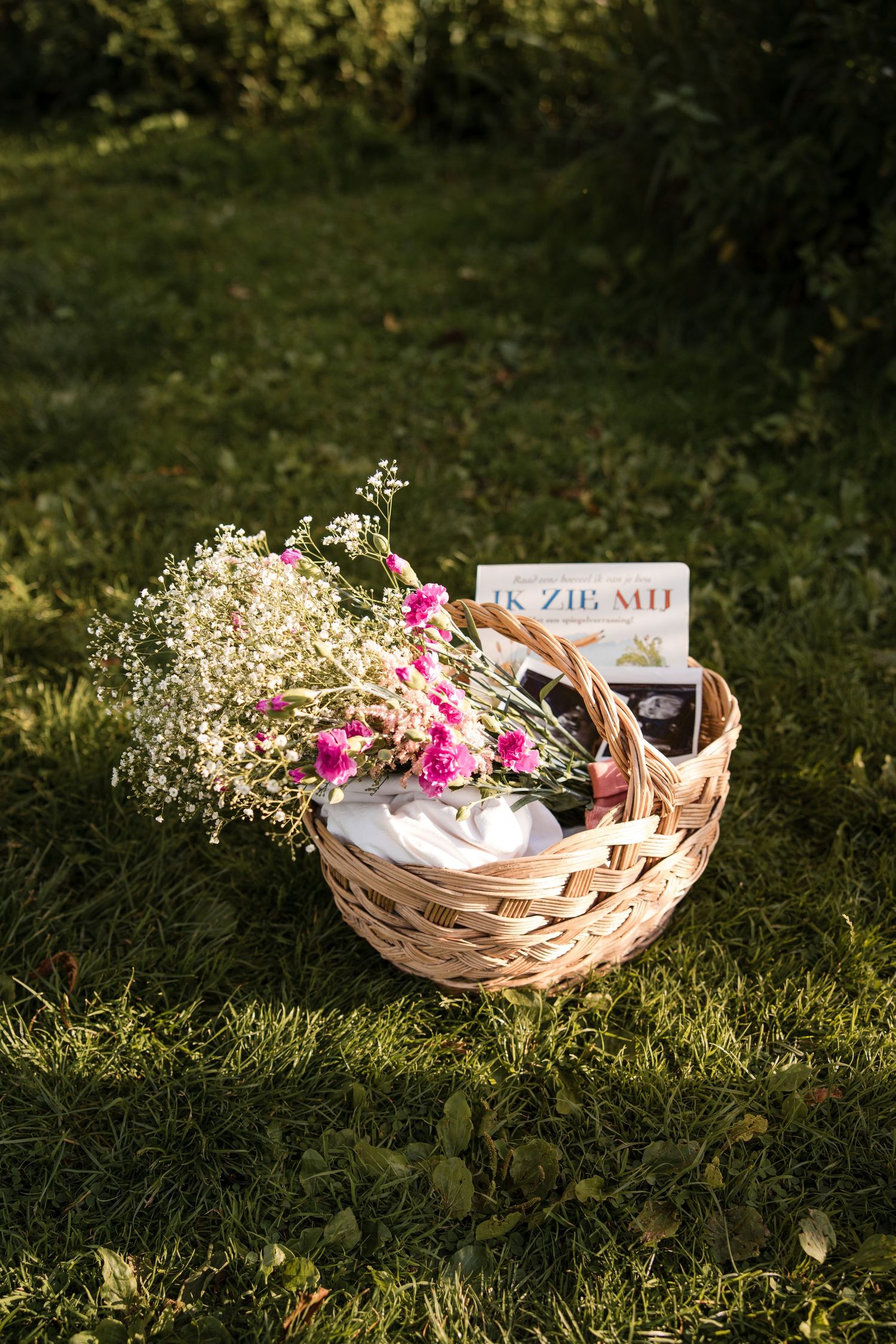 A wicker basket filled with pink flowers and baby's breath sits on green grass in sunlight, inside baby books and ultrasound baby pictures
