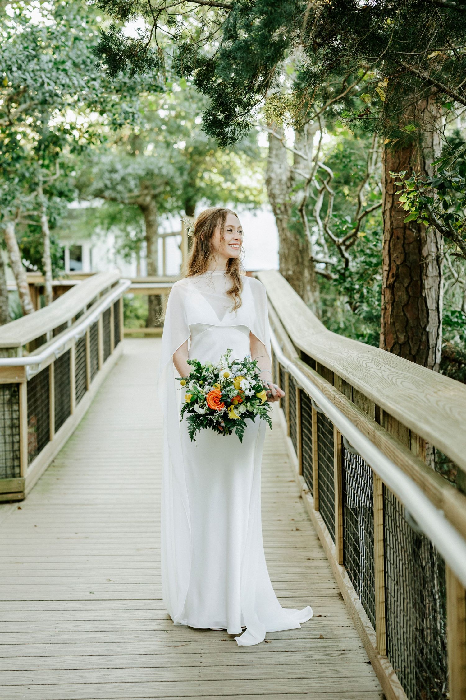 A flowing white wedding dress twirls gracefully on a wooden boardwalk surrounded by lush green trees.