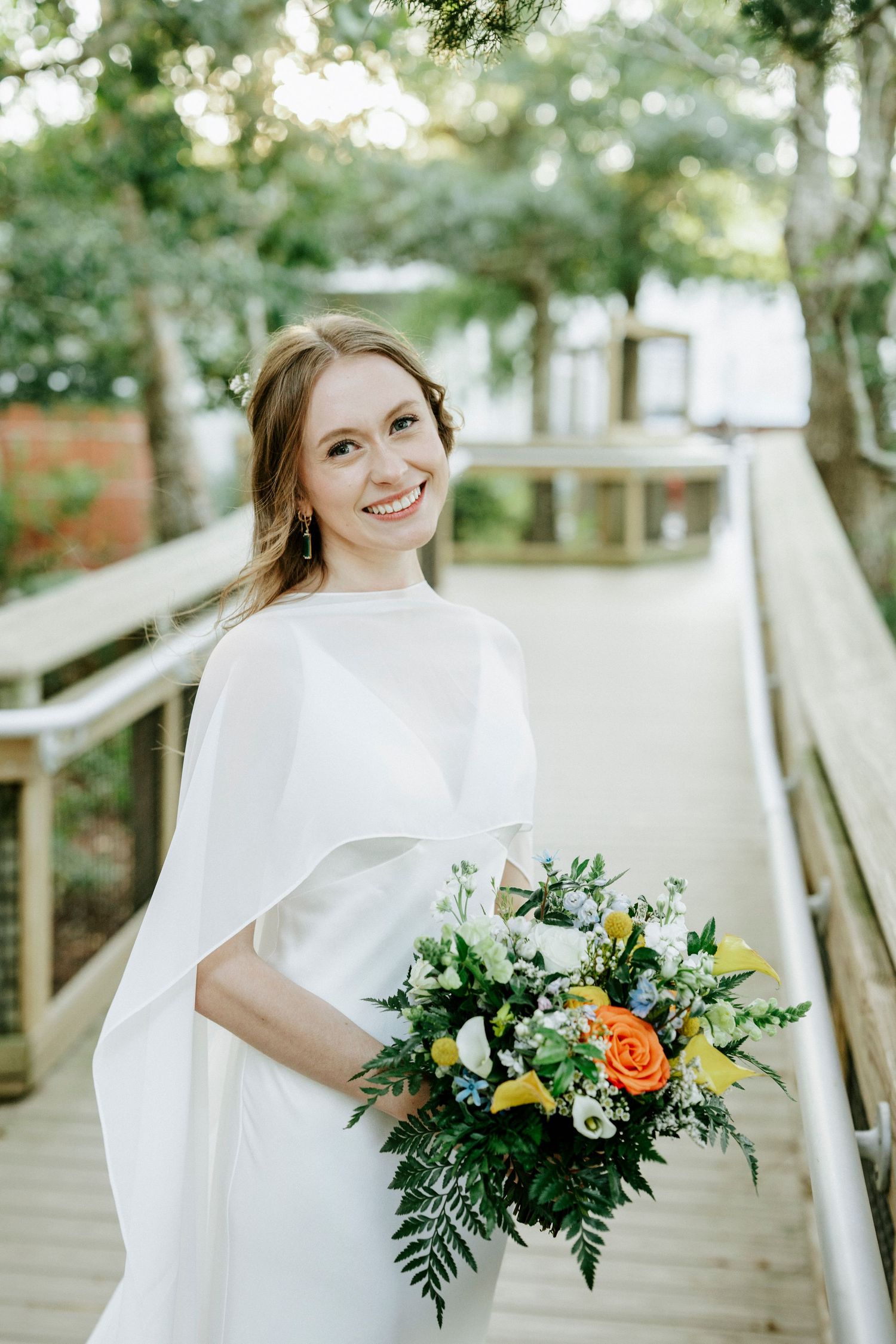 A bride in a flowing white dress stands on a wooden walkway holding a colorful bouquet with orange roses and greenery.