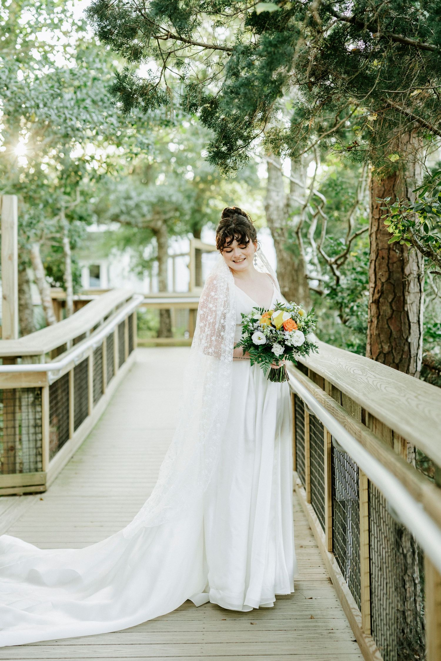 Elegant wedding dress with flowing train photographed on wooden boardwalk surrounded by trees.