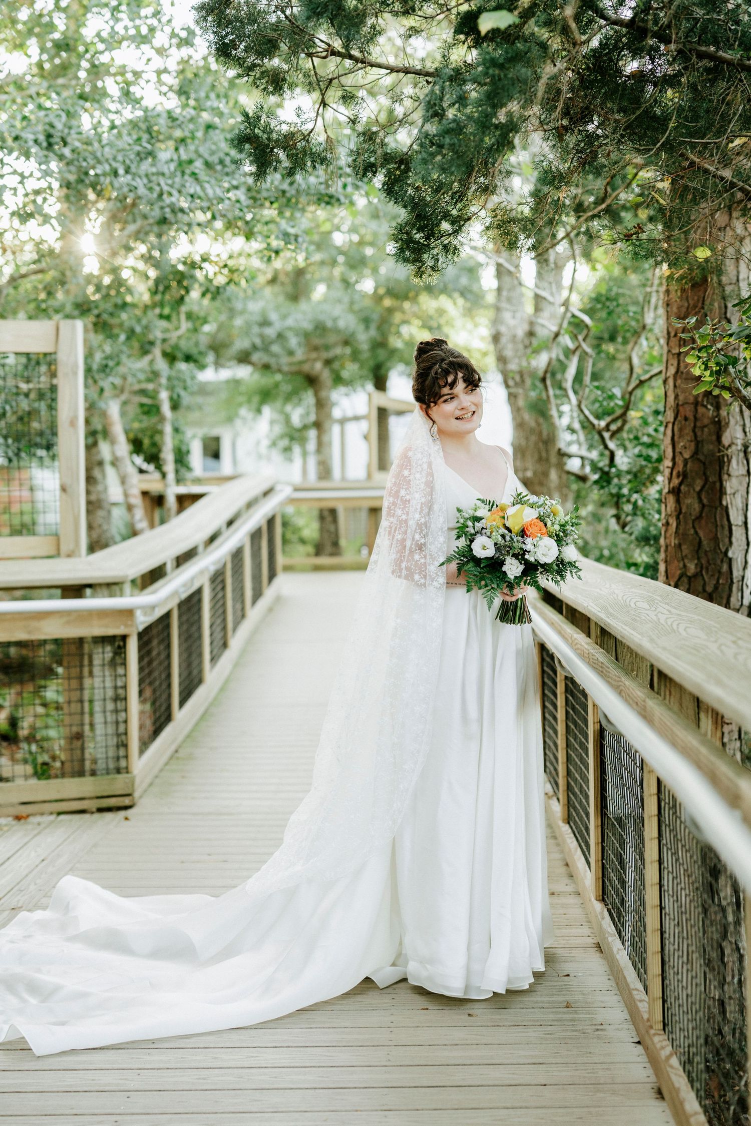 White wedding gown with long train captured on rustic wooden walkway amidst green foliage.